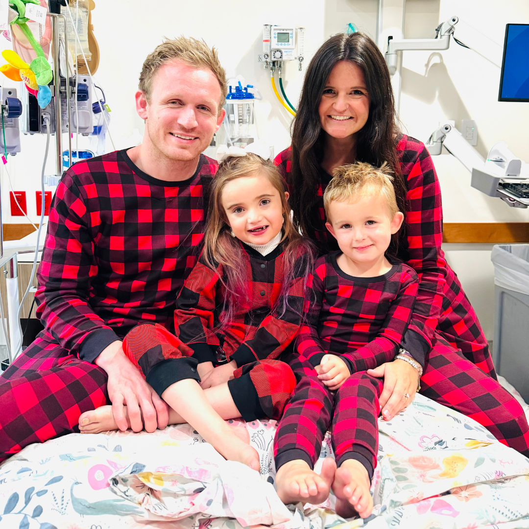 A family sitting in a hospital bed. The young girl in the middle is wearing a red and black buffalo plaid hospital gown alternative. The family all has matching pajamas.