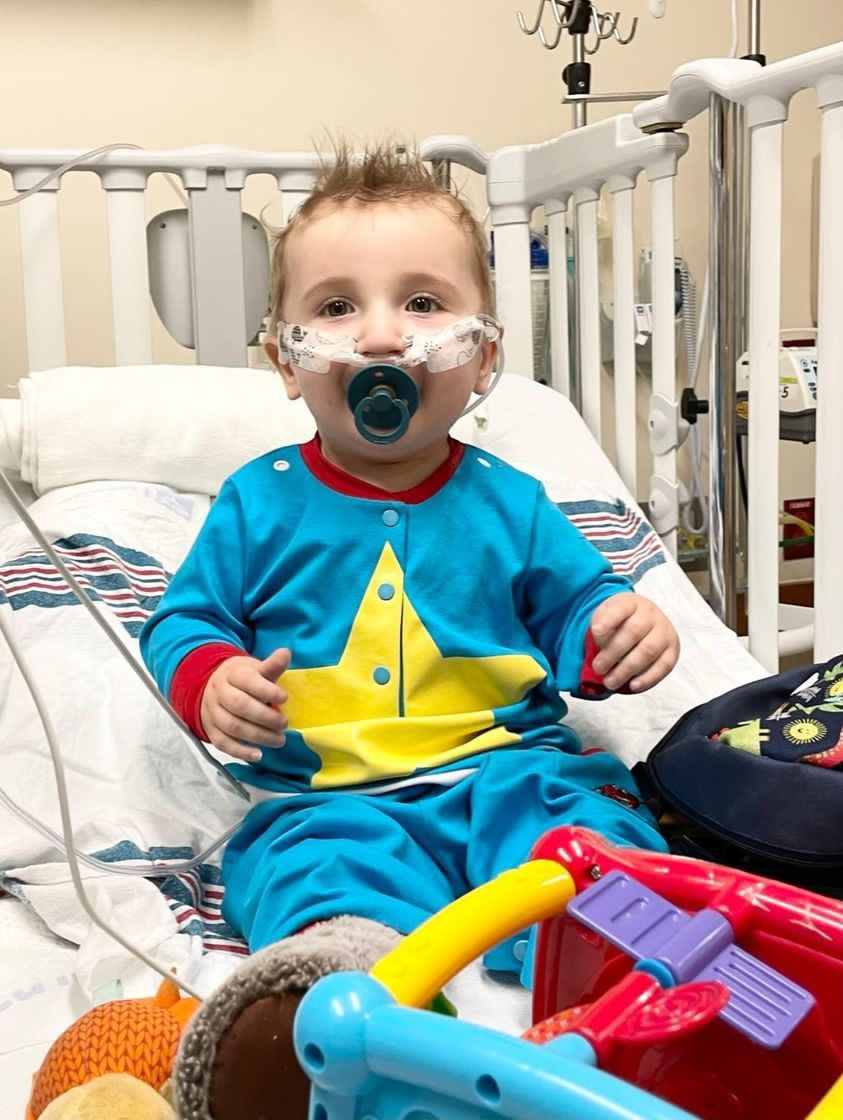 A young patient in a hospital bed with a pacifier. The patient is hooked up to medical equipment. They are wearing a super hero hospital gown alternative. It has a bight yellow star on the front.