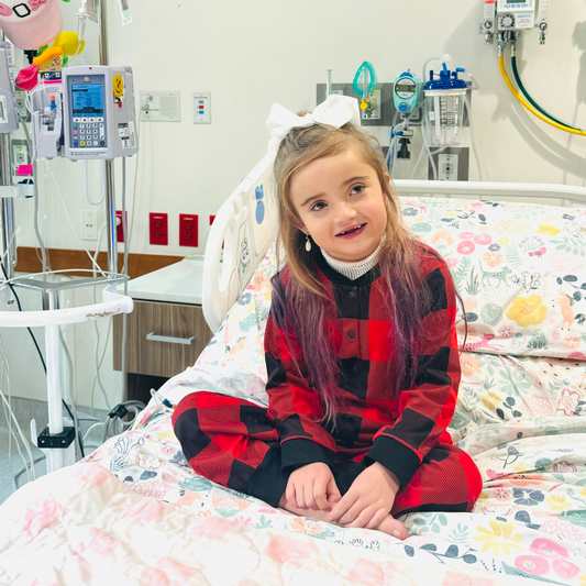 A young girl smiling in a hospital bed. She has a big white bow, and is wearing a buffalo plaid hospital gown alternative. 